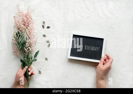 Testo Happy Birthday su lavagna, mani tenere tavola e fiori selvatici in mani su tavolo tessile. Naturale sfondo estivo con Prachtspiere, Astilbe Foto Stock