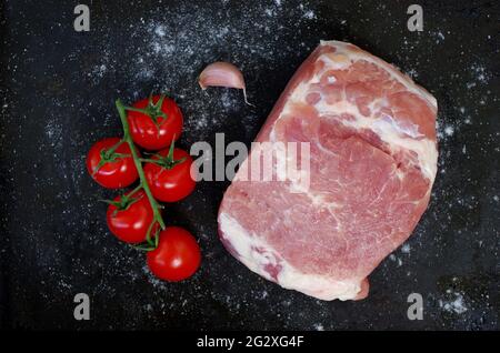 Pezzo di maiale crudo con pomodori rossi, aglio fresco e sale su fondo nero. Grill party e concetto di cibo. Pezzo grande di carne cruda. Foto Stock