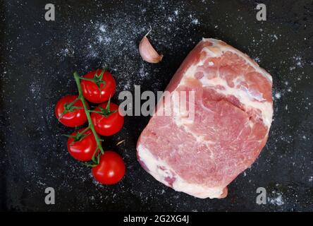 Pezzo di maiale crudo con pomodori rossi, aglio fresco e sale su fondo nero. Grill party e concetto di cibo. Pezzo grande di carne cruda. Foto Stock