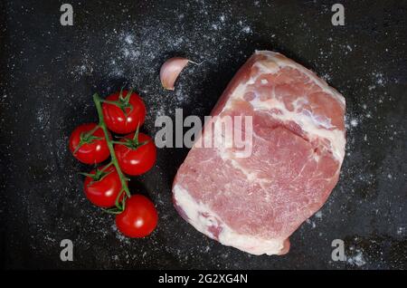 Pezzo di maiale crudo con pomodori rossi, aglio fresco e sale su fondo nero. Grill party e concetto di cibo. Pezzo grande di carne cruda. Foto Stock