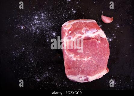 Pezzo di maiale crudo con aglio fresco rosso e sale su fondo nero. Vista dall'alto di un pezzo di maiale disossato fresco, un pezzo di carne grande. Concetto di cibo. Foto Stock