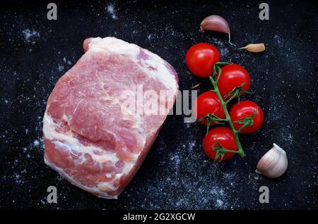 Pezzo di maiale crudo con pomodori rossi, aglio fresco e sale su fondo nero. Grill party e concetto di cibo. Pezzo grande di carne cruda. Foto Stock