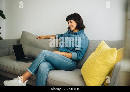 Donna sorridente seduta sul divano nella lobby con le gambe incrociate. Donna sicura seduta nella lobby che guarda via e sorridente. Foto Stock
