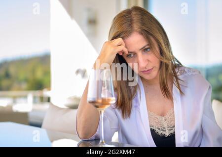 Donna depressa seduta pensando profondamente con un bicchiere di vino rosso davanti a lei appoggiando la fronte sulla sua mano fissando al tavolo Foto Stock