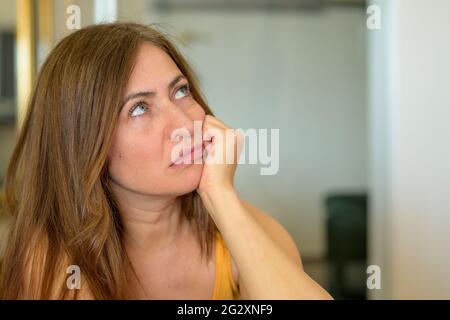 Donna preoccupata e travagliata seduta profonda nel pensiero appoggiando il mento sulla sua mano guardando in su con un'espressione contemplativa sommaria Foto Stock