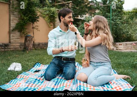 Vacanze estive, persone, romanticismo, uomo e donna si nutrono a vicenda fragole mentre beve frizzante e godendo del tempo insieme a casa Foto Stock