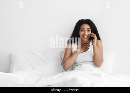 Ottime notizie mattutine. Donna afro-americana felice che parla su smartphone mentre si siede a letto in camera da letto leggera, spazio vuoto Foto Stock