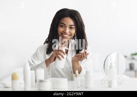 Donna nera sorridente usando il prodotto o la lozione di pulizia, pulendo il suo viso o la pelle idratante, seduto in camera da letto Foto Stock