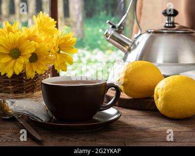 Tazza di tè con limoni e fiori su un tavolo di legno vicino a una finestra con sfondo rurale Foto Stock