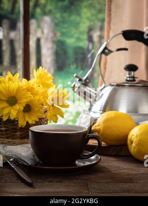 Tazza di tè con limoni e fiori su un tavolo di legno vicino a una finestra con sfondo rurale Foto Stock