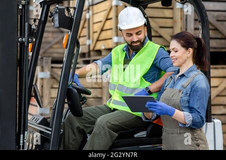 Uomo sopportato millenario in casco in carrello elevatore a forche, discutendo ordine con donna sorridente Foto Stock