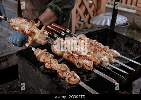 Lo chef prepara il kebab di shish di pollo. Cucina turca di strada. Carne cotta al fuoco, barbecue party nel cortile. Le mani maschili nei guanti tengono il matassa di acciaio Foto Stock