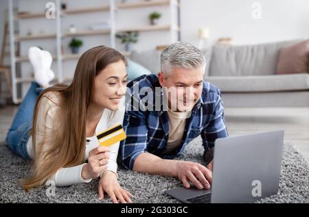 Allegro coniugi maturi che ordinano le merci on-line con PC portatile e carta di credito, sdraiati sul pavimento a casa Foto Stock