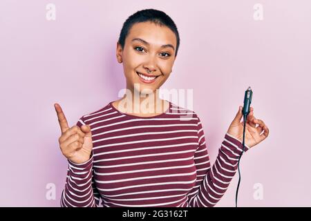 Bella donna ispanica con i capelli corti che tengono l'adattatore del caricatore usb per l'automobile sorridente felice che punta con la mano e il dito al lato Foto Stock