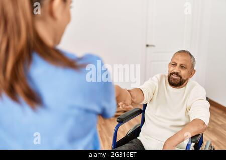 Uomo anziano seduto su sedia a rotelle e infermiera geriatrica che fa accordo di scuotimento della mano, ringraziando per l'aiuto Foto Stock