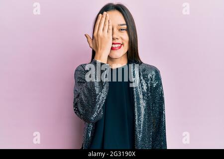 Giovane ragazza ispanica con giacca da festa che copre un occhio con la mano, sorriso fiducioso sul viso e sorpresa emozione. Foto Stock