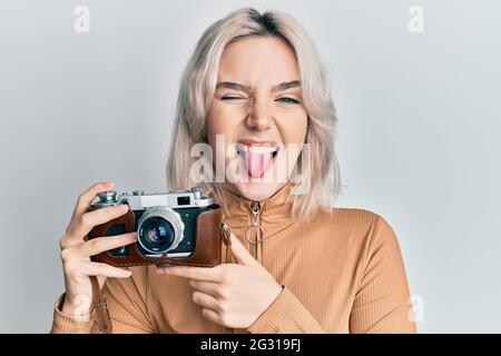 Giovane ragazza bionda che tiene vintage macchina fotografica attaccando lingua fuori felice con l'espressione divertente. Foto Stock