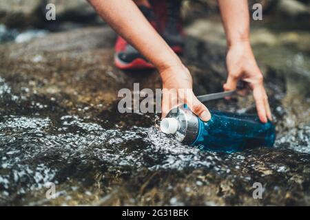 Foto ravvicinata delle mani femminili che riempiono un fresco flusso d'acqua di montagna nella bottiglia turistica di plastica mentre l'acqua si rompe durante l'escursione turistica Foto Stock