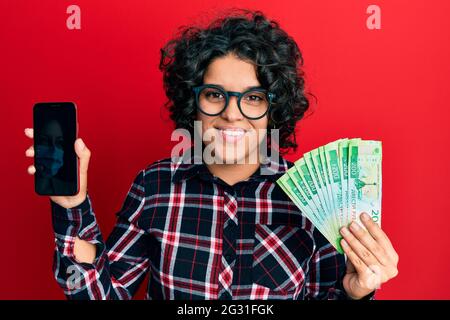 Giovane donna ispanica con capelli ricci che mostra lo schermo dello smartphone tenendo 200 ruble sorridente con un sorriso felice e fresco sul viso. Mostrando i denti. Foto Stock