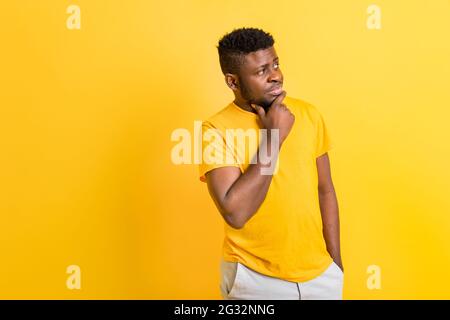 Uomo afroamericano pensivo in t-shirt gialla che tiene la mano sul mento guardando lo spazio della copia, uomo nero dubbio pensieroso con il pensiero del volto incerto Foto Stock