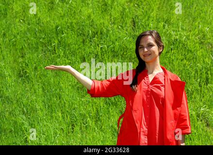 Una ragazza sorridente in abiti rossi su sfondo di erba verde, un gesto della mano sul lato. Una giovane, fiduciosa donna sullo sfondo del prato verde ha alzato la mano a lato. Spazio per la pubblicità Foto Stock