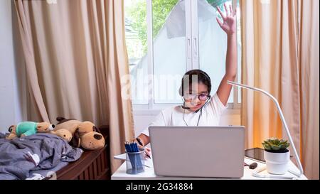 Ragazza asiatica in cuffie sedersi a scrivania studio on-line sul computer portatile. Kid Wear auricolare scrivere a mano in notebook imparare utilizzando lezioni di Internet sulla quarantena. G Foto Stock