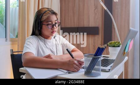 Ragazza asiatica in cuffie sedersi a scrivania studio on-line sul computer portatile. Kid Wear auricolare scrivere a mano in notebook imparare utilizzando lezioni di Internet sulla quarantena. G Foto Stock