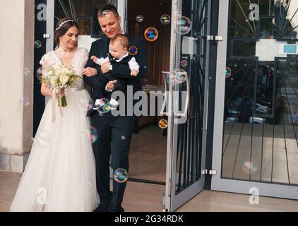 Appena sposato coppia con appena battezzato bambino uscendo dalla chiesa con bolle di sapone di fronte a loro Foto Stock