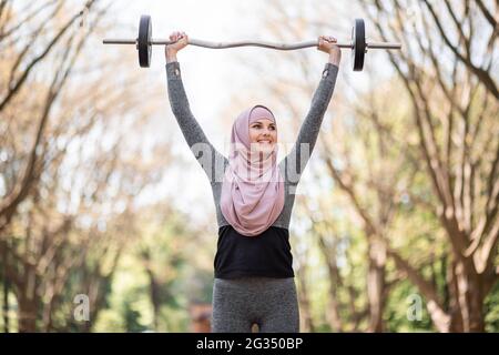 Donna sportiva in abbigliamento sportivo e hijab che tiene pesante barbell sulla testa mentre si trova al parco. Buona formazione femminile musulmana durante il tempo libero all'aperto. Foto Stock