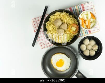 Noodle calde servite con uova e pomodori freschi con polpette su una ciotola nera e sottaceto su una ciotola bianca e uova su fondo bianco Foto Stock