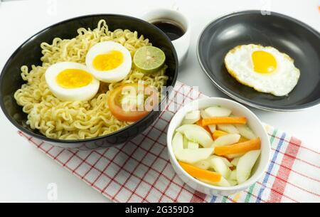 Noodle calde servite con uova e pomodori freschi con polpette su una ciotola nera e sottaceto su una ciotola bianca e uova su fondo bianco Foto Stock