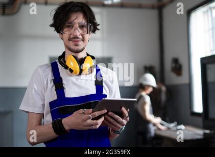 Ritratto di un giovane lavoratore con un tablet in piedi all'interno in fabbrica guardando la fotocamera. Foto Stock