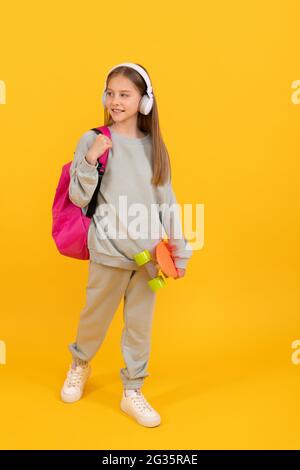 felice ragazzino adolescente con zaino scuola e penny bordo ascoltare musica in cuffia, tempo di scuola. Foto Stock