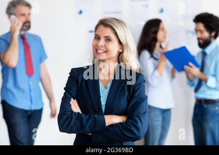 Bionda donna d'affari tedesca con team internazionale in background presso l'ufficio di società Foto Stock
