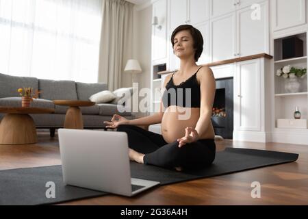 Donna incinta consapevole che pratica yoga online, meditando, utilizzando il computer portatile Foto Stock