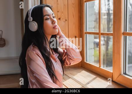 sognante giovane donna asiatica in pigiami di seta in piedi vicino alla finestra con soglia con le cuffie e tenendo la mano vicino alla testa a casa Foto Stock