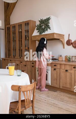 giovane donna asiatica in pajama di seta rosa e cuffie che cucinano la colazione vicino al tavolo con succo d'arancia in cucina Foto Stock