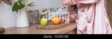 vista tagliata di giovane donna in pigiami di seta rosa che tagliano frutta in cucina, striscione Foto Stock