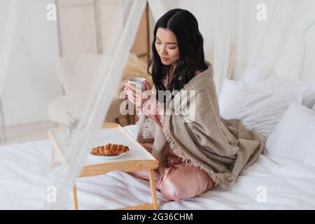 giovane donna asiatica in pigiama di seta e coperta seduta sul letto con gambe incrociate e facendo colazione in camera da letto Foto Stock