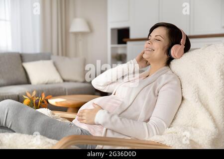 Donna incinta serena e sorridente in cuffia che ascolta la musica preferita Foto Stock