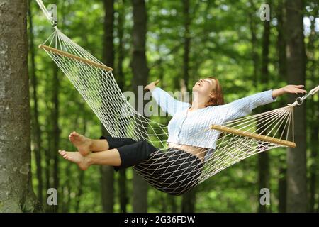 Donna felice che oscilla su amaca che allunga le braccia in una foresta Foto Stock