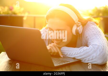 Happy lady con cuffie wireless al tramonto che controlla il computer portatile in un balcone Foto Stock