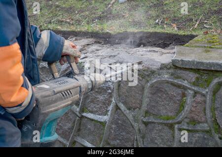 Lavorare con il martello rotante per rompere sottili piatti di cemento di cordoli, le mani gualcate di uomo vestito in tute tengono il martello pneumatico da entrambi i manici, polvere A. Foto Stock