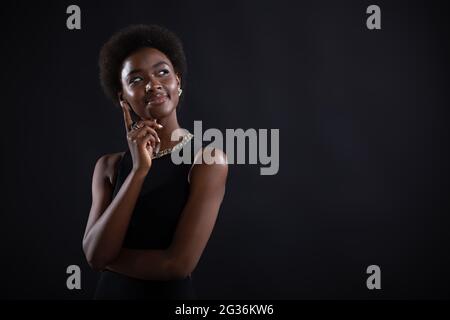 Giovane donna nera cercando di trovare la soluzione migliore e la risposta di ricerca. Donna afro-americana che ha un'espressione dubbia guardando il lato lontano e tenendo il mento con la mano su sfondo nero. Foto Stock