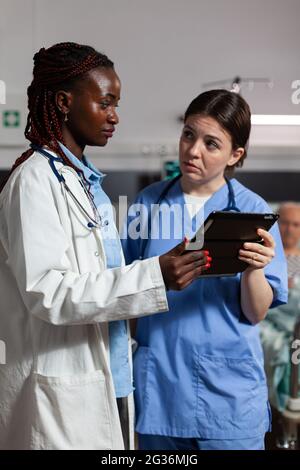 Infermiera medica che chiede consiglio dal medico afroamericano, tenendo il pc della tavoletta nella stanza dell'ospedale, circa il trattamento per l'uomo anziano malato, malato, malato che posa nel letto. Foto Stock