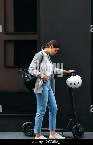 Foto a tutta lunghezza di giovane donna d'affari in piedi in un edificio con scooter elettrico a spinta verso il basso Foto Stock