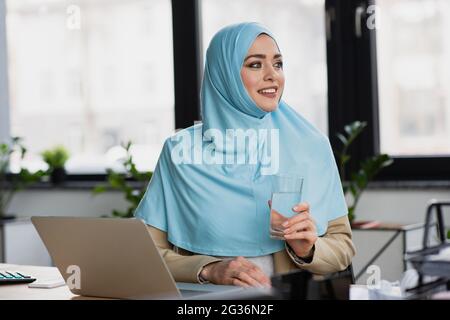 donna d'affari musulmana di successo con un bicchiere d'acqua che guarda lontano vicino al computer portatile in ufficio Foto Stock