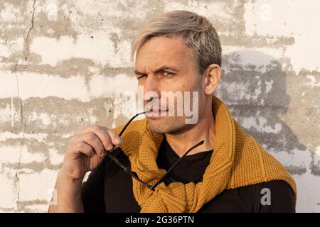 Ritratto affascinante uomo biondo di mezza età dall'aspetto modella con occhiali in mano davanti a una parete di mattoni bianchi. Bel modello maschile maturo in casual Foto Stock