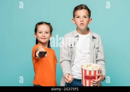 ragazza che tiene il telecomando vicino al ragazzo con la benna popcorn isolata in blu Foto Stock