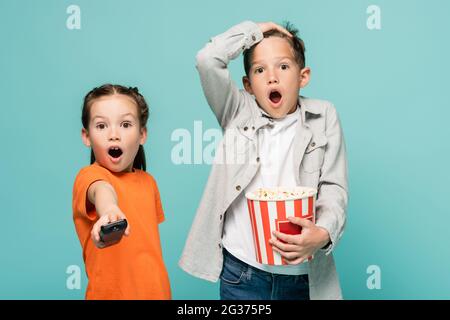 ragazza scioccata che tiene il telecomando vicino al ragazzo con la benna popcorn isolata in blu Foto Stock